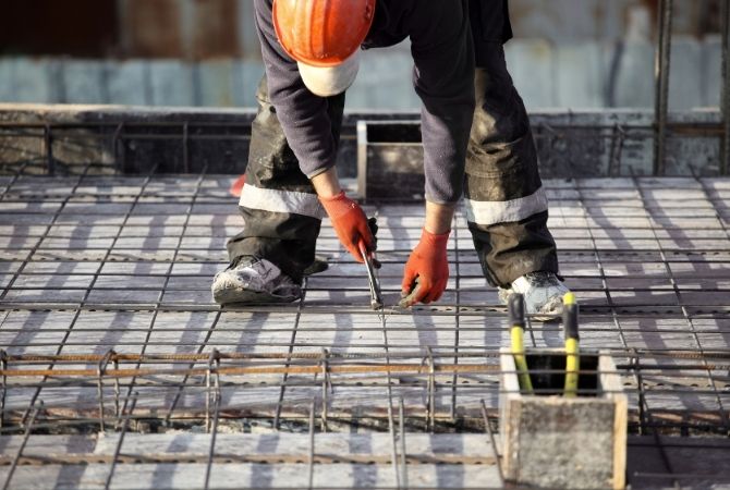 Bauarbeiter mit orangefarbenem Schutzhelm und Handschuhen befestigt Bewehrungseisen auf einer Baustelle im Nordburgenland für einen Betonboden.